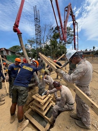 U.S. Navy Seabees and Marines are engaged in a concrete pouring operation at an outdoor construction site, likely in a tropical or subtropical region based on the vegetation. Workers are managing a concrete pump hose directed into reinforced steel column forms while others secure wooden formwork. The scene depicts a military construction/humanitarian engineering project.