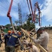 U.S. Navy Seabees and Marines are engaged in a concrete pouring operation at an outdoor construction site, likely in a tropical or subtropical region based on the vegetation. Workers are managing a concrete pump hose directed into reinforced steel column forms while others secure wooden formwork. The scene depicts a military construction/humanitarian engineering project.