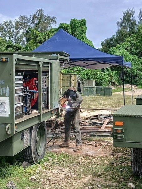 A welder in full protective gear is performing welding work between two military-green vehicles or trailers in an outdoor field setting. A blue pop-up canopy tent is erected nearby, and an open equipment panel on the green trailer reveals red and silver mechanical components. The surrounding area contains scattered debris, wooden planks, and military-style storage containers in the background.