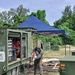 A welder in full protective gear is performing welding work between two military-green vehicles or trailers in an outdoor field setting. A blue pop-up canopy tent is erected nearby, and an open equipment panel on the green trailer reveals red and silver mechanical components. The surrounding area contains scattered debris, wooden planks, and military-style storage containers in the background.