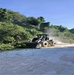 A front-end loader/wheel loader is actively clearing vegetation and debris from the edge of a paved road. The machine appears to be pushing cut branches and foliage off the road surface, with dust visible in the air. At least one person is visible in the background observing or supervising the operation.