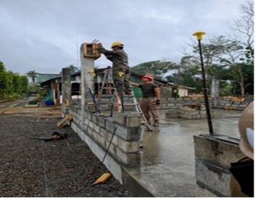 Two workers are engaged in masonry and construction work on a partially built concrete block wall structure. One worker wearing a yellow hard hat is elevated on a ladder working on a concrete column, while a second worker in a red hard hat stands on the concrete slab below. The setting appears to be a tropical or subtropical environment based on the vegetation visible in the background.