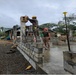 Two workers are engaged in masonry and construction work on a partially built concrete block wall structure. One worker wearing a yellow hard hat is elevated on a ladder working on a concrete column, while a second worker in a red hard hat stands on the concrete slab below. The setting appears to be a tropical or subtropical environment based on the vegetation visible in the background.