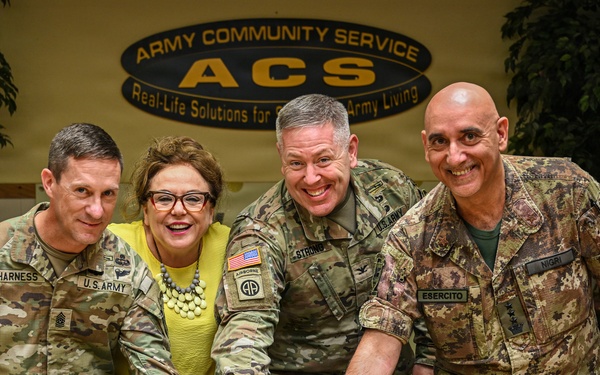 Four individuals — three in U.S. Army uniforms and one in Italian Army (Esercito) uniform — are cutting a decorated anniversary cake at an Army Community Service (ACS) event. The cake displays the number '61' or '67' in candles, suggesting a milestone anniversary celebration. The setting is inside an ACS facility with the organization's logo and motto visible in the background.