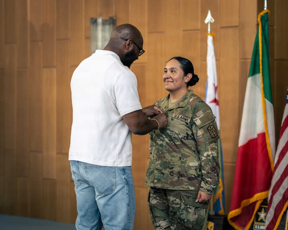 A civilian man in a white shirt and jeans is pinning something onto the chest of a U.S. Army soldier in OCP (Operational Camouflage Pattern) uniform during what appears to be a military promotion or award ceremony. The soldier is smiling and standing at attention in front of several flags. The setting appears to be an official indoor military venue.
