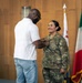 A civilian man in a white shirt and jeans is pinning something onto the chest of a U.S. Army soldier in OCP (Operational Camouflage Pattern) uniform during what appears to be a military promotion or award ceremony. The soldier is smiling and standing at attention in front of several flags. The setting appears to be an official indoor military venue.