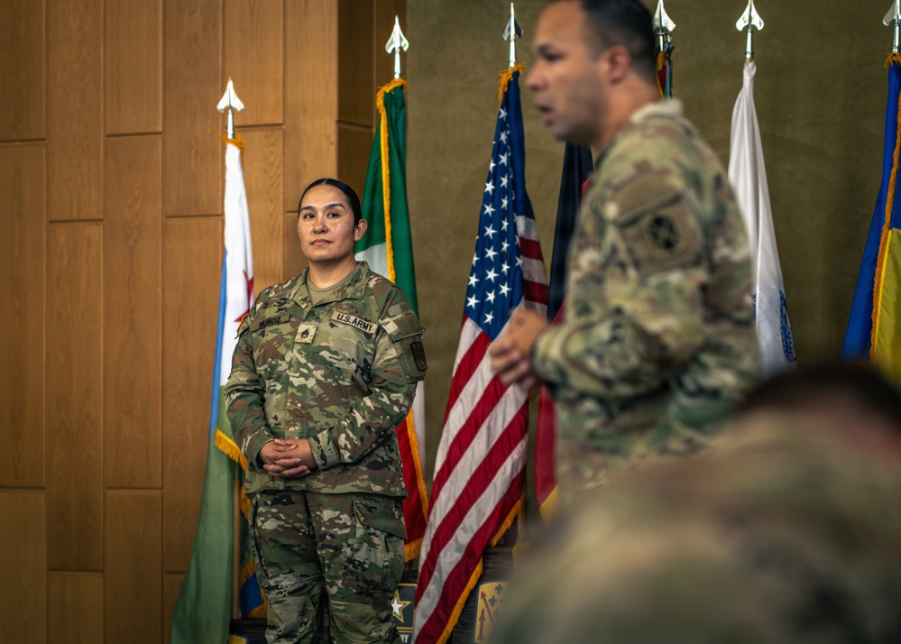 A U.S. Army ceremony is taking place in a formal indoor setting with multiple flags displayed in the background. A female soldier with the name tape 'MUNOZ' and Staff Sergeant rank insignia stands at attention while a male soldier in the foreground appears to be speaking. The setting suggests an official military ceremony such as a promotion, award, or change of responsibility.