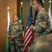 A U.S. Army ceremony is taking place in a formal indoor setting with multiple flags displayed in the background. A female soldier with the name tape 'MUNOZ' and Staff Sergeant rank insignia stands at attention while a male soldier in the foreground appears to be speaking. The setting suggests an official military ceremony such as a promotion, award, or change of responsibility.