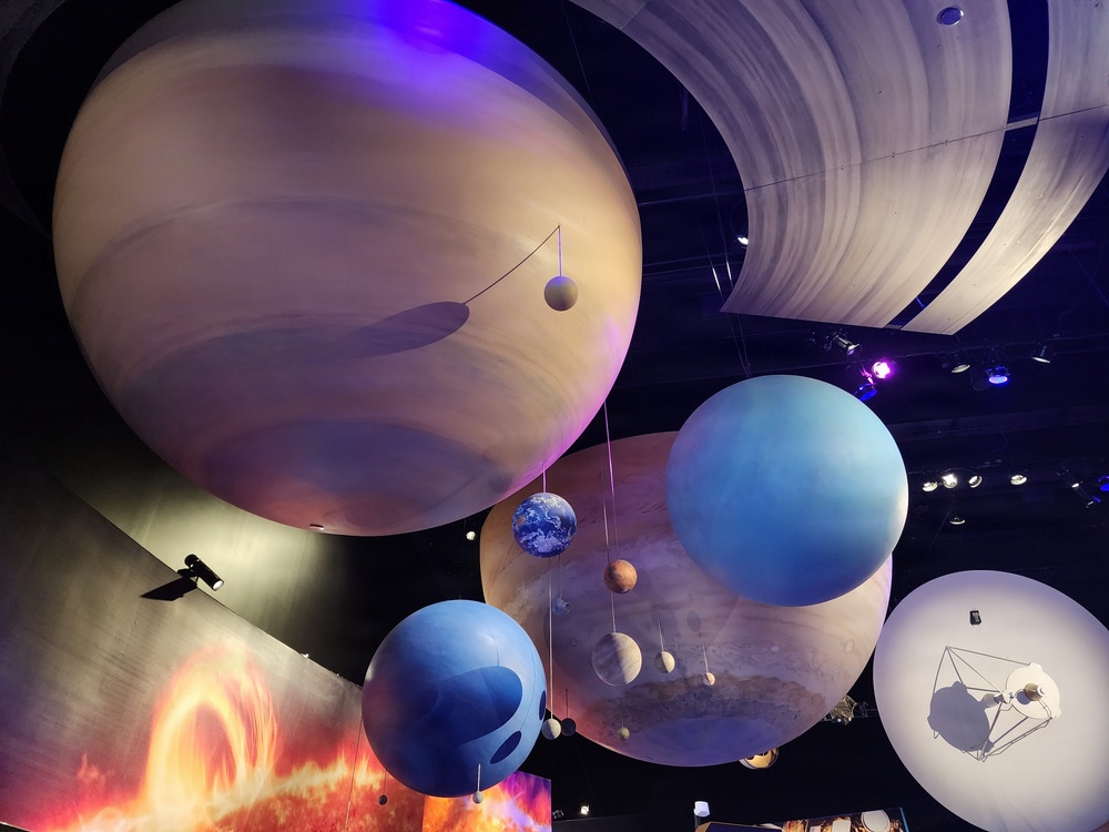This image shows a large-scale solar system model display, likely inside a science museum or planetarium. Oversized hanging spheres represent planets including Earth, Jupiter, Neptune, Uranus, and Saturn (with visible rings), suspended from a dark ceiling. The exhibit also features imagery of the Sun's surface and what appears to be a spacecraft model on the right side.