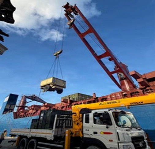 A mobile crane truck is actively lifting a piece of heavy equipment or machinery at what appears to be a port or dockyard. The crane, branded 'SOOSAN', is mounted on a white and yellow truck and is hoisting a yellow and grey load suspended by cables. A large cargo ship loaded with containers is visible in the background.