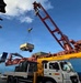 A mobile crane truck is actively lifting a piece of heavy equipment or machinery at what appears to be a port or dockyard. The crane, branded 'SOOSAN', is mounted on a white and yellow truck and is hoisting a yellow and grey load suspended by cables. A large cargo ship loaded with containers is visible in the background.