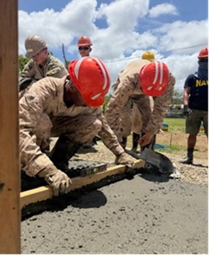 Military personnel in camouflage uniforms and red hard hats are working together to pour and finish a concrete slab outdoors. The team appears to include Marines and at least one Navy member, performing construction work in what appears to be a humanitarian or engineering mission. The work is being conducted in a sunny, tropical-looking environment.