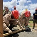 Military personnel in camouflage uniforms and red hard hats are working together to pour and finish a concrete slab outdoors. The team appears to include Marines and at least one Navy member, performing construction work in what appears to be a humanitarian or engineering mission. The work is being conducted in a sunny, tropical-looking environment.