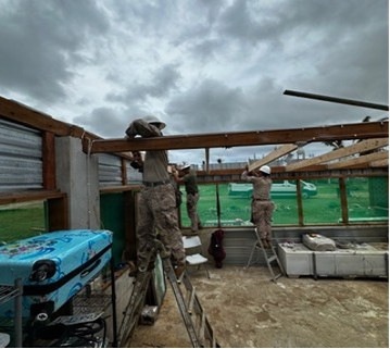 Three individuals wearing military-style camouflage uniforms and white hard hats are performing construction or repair work on a partially roofless structure. They are standing on ladders and handling wooden beams, suggesting roof reconstruction or storm damage repair. The overcast, stormy sky and damaged structure indicate this may be post-disaster recovery work.