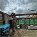 Three individuals wearing military-style camouflage uniforms and white hard hats are performing construction or repair work on a partially roofless structure. They are standing on ladders and handling wooden beams, suggesting roof reconstruction or storm damage repair. The overcast, stormy sky and damaged structure indicate this may be post-disaster recovery work.
