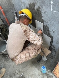 A worker wearing a yellow hard hat and camouflage pants is crouching down and applying ceramic tiles to a concrete wall using mortar/adhesive. The tiles are being set in a grid pattern on the lower portion of the wall. Various tiling tools and materials are visible around the worker.