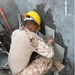 A worker wearing a yellow hard hat and camouflage pants is crouching down and applying ceramic tiles to a concrete wall using mortar/adhesive. The tiles are being set in a grid pattern on the lower portion of the wall. Various tiling tools and materials are visible around the worker.