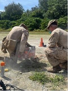 Two military personnel in desert camouflage uniforms are crouched over a piece of orange equipment on a gravel surface, with smoke or dust visible around the work area. One individual is wearing a face mask and glasses, and both appear to be engaged in a hands-on technical or engineering task. An orange traffic cone, marker tubes, and cables are visible nearby.