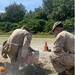 Two military personnel in desert camouflage uniforms are crouched over a piece of orange equipment on a gravel surface, with smoke or dust visible around the work area. One individual is wearing a face mask and glasses, and both appear to be engaged in a hands-on technical or engineering task. An orange traffic cone, marker tubes, and cables are visible nearby.