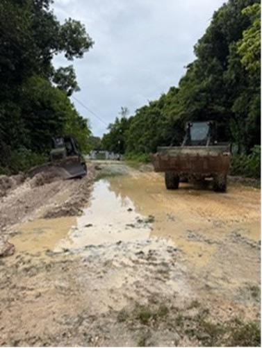 The image shows an unpaved dirt road under construction or maintenance, with standing water pooled along the center of the roadway. Two pieces of heavy construction equipment — a bulldozer on the left and a dump truck on the right — are visible operating along the corridor. The road is flanked by dense tropical or subtropical vegetation on both sides under an overcast sky.