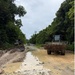 The image shows an unpaved dirt road under construction or maintenance, with standing water pooled along the center of the roadway. Two pieces of heavy construction equipment — a bulldozer on the left and a dump truck on the right — are visible operating along the corridor. The road is flanked by dense tropical or subtropical vegetation on both sides under an overcast sky.