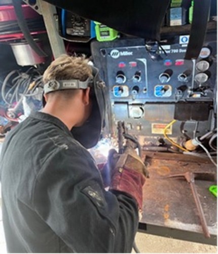 A welder is actively performing a welding operation on a metal workpiece, with visible sparks and arc light. The worker is equipped with a welding helmet pushed up on their head and heavy-duty leather welding gloves. A Miller brand welding machine (appears to be a Miller 700 series) is visible in the background as the primary equipment.