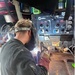 A welder is actively performing a welding operation on a metal workpiece, with visible sparks and arc light. The worker is equipped with a welding helmet pushed up on their head and heavy-duty leather welding gloves. A Miller brand welding machine (appears to be a Miller 700 series) is visible in the background as the primary equipment.