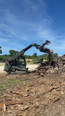 A compact track loader, branded JCB, is actively working to sort or move a large pile of debris consisting of broken wood, branches, and timber. The machine is equipped with a hydraulic grapple attachment, which is raised and gripping material from the pile. The setting appears to be an outdoor debris clearing or land clearing operation under a clear blue sky.