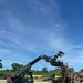 A compact track loader, branded JCB, is actively working to sort or move a large pile of debris consisting of broken wood, branches, and timber. The machine is equipped with a hydraulic grapple attachment, which is raised and gripping material from the pile. The setting appears to be an outdoor debris clearing or land clearing operation under a clear blue sky.
