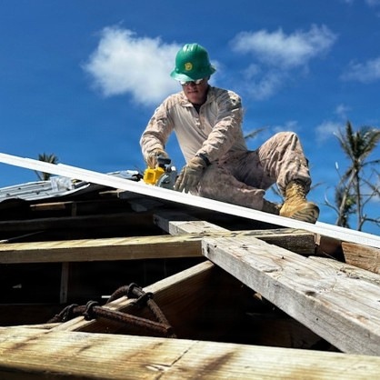A worker in military-style camouflage uniform and green hard hat is performing roofing or construction/repair work on a damaged wooden structure. The person is crouching on roof framing while operating a power tool, likely a circular saw, on a sheet of roofing material. The surrounding environment shows damaged or stripped palm trees, suggesting post-storm disaster recovery work.