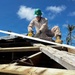 A worker in military-style camouflage uniform and green hard hat is performing roofing or construction/repair work on a damaged wooden structure. The person is crouching on roof framing while operating a power tool, likely a circular saw, on a sheet of roofing material. The surrounding environment shows damaged or stripped palm trees, suggesting post-storm disaster recovery work.