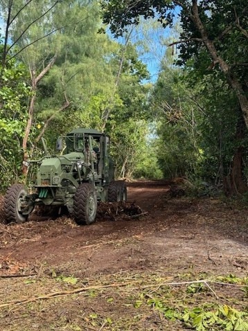 A military-green painted motor grader or road grading vehicle is actively clearing and grading a dirt trail through a dense tropical forest. The machine appears to be pushing debris and leveling the red-brown soil surface along a tree-lined path. The surrounding vegetation is lush and tropical, consistent with a Pacific island or similar tropical environment.