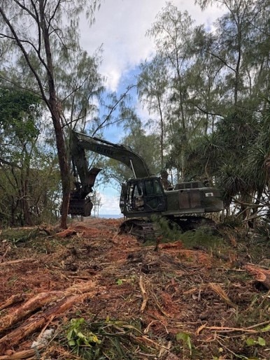 A tracked excavator, appearing to be a John Deere model, is actively engaged in land clearing or vegetation removal operations in a forested area. The machine is using a grapple or shear attachment to grip and remove trees. The foreground shows significant debris including felled branches, wood chips, and disturbed red-brown soil, indicating active clearing work.