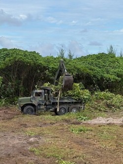 A military-style olive drab dump truck equipped with a crane/loader arm is engaged in land clearing operations at the edge of a dense tropical or subtropical tree line. The vehicle is actively loading or moving vegetation debris into its cargo bed. The foreground shows recently cleared bare earth, indicating active ground preparation work.