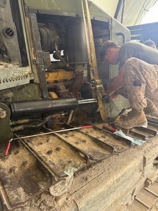 A person in military camouflage uniform is performing maintenance or repair work on a large military vehicle, likely a tracked armored vehicle or heavy equipment. The work is being conducted inside what appears to be a maintenance bay or tent facility. Multiple hand tools are visible on the vehicle's deck surface alongside mechanical components.