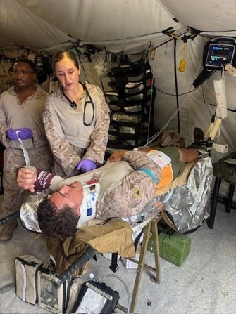 A military medical treatment scene inside a field hospital tent, showing personnel in camouflage uniforms providing care to a casualty on a litter. A medical provider wearing purple gloves and a stethoscope is actively treating the patient, who is wrapped in a mylar/emergency blanket and fitted with a cervical collar. Multiple pieces of medical monitoring equipment are visible, suggesting this is a forward surgical or Role 2 medical facility.