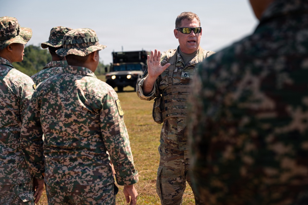 A soldier in US Army MultiCam (OCP) uniform with tactical vest and sunglasses appears to be briefing or instructing a group of soldiers wearing a different digital camouflage pattern, likely from a partner nation. The setting is an outdoor field environment with military vehicles visible in the background. The interaction suggests a joint training exercise or multinational military cooperation event.