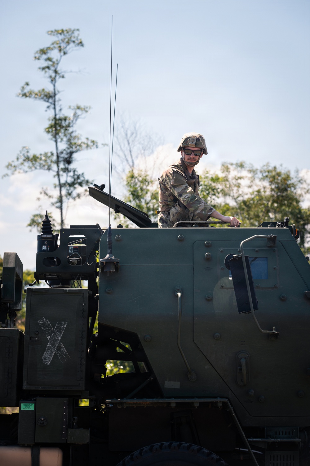A soldier in camouflage uniform and combat helmet is positioned atop a large olive-drab military vehicle, likely an armored or artillery system. The soldier is wearing sunglasses and appears to be operating or inspecting the vehicle from an open hatch. The setting is an outdoor environment with trees and a partly cloudy sky in the background.