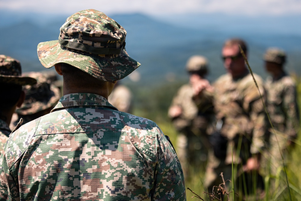 The image shows a group of military personnel in an outdoor field setting with mountainous terrain in the background. Soldiers wearing different camouflage patterns are gathered together, suggesting a multinational training exercise or briefing. The foreground subject is in sharp focus while background figures are intentionally blurred.