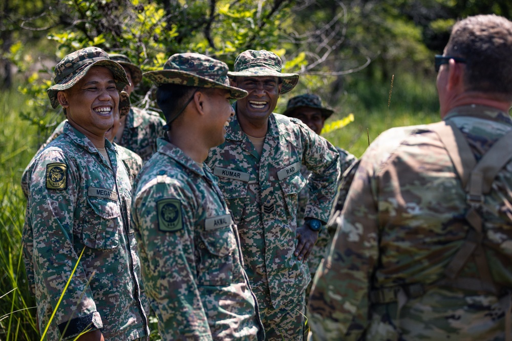 A group of military personnel in digital camouflage uniforms are engaged in a friendly interaction outdoors in a vegetated area. The soldiers wearing green digital camouflage with wide-brimmed hats appear to be from a Southeast Asian or Pacific nation's armed forces, interacting with a soldier in MultiCam/OCP pattern uniform facing away from the camera. The atmosphere is relaxed and positive, with visible smiling and laughter among the group.