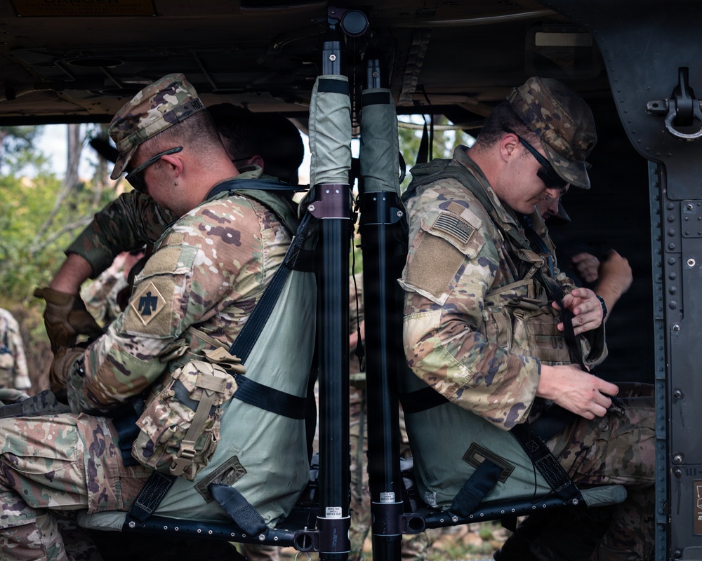 Two U.S. Army soldiers in Operational Camouflage Pattern (OCP) uniforms are seated inside or alongside a military vehicle, appearing to adjust their gear or equipment. Both wear tactical vests, patrol caps, and one wears sunglasses. A tripod-mounted piece of equipment is visible between them, likely a mortar or survey instrument.