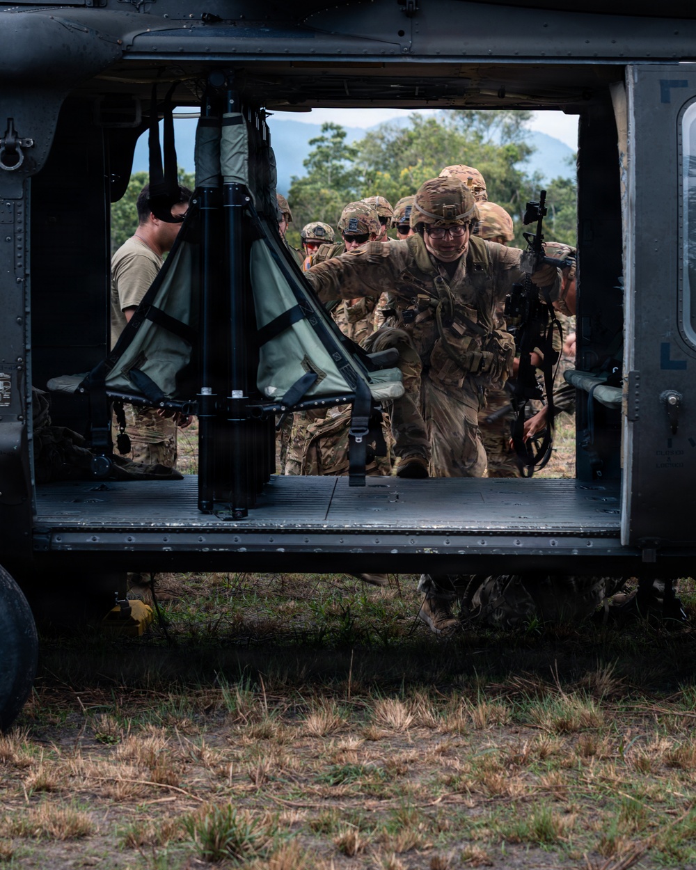 Military personnel in full combat gear are boarding or loading into a helicopter, visible through the open side door of what appears to be a UH-60 Black Hawk helicopter. Soldiers wearing camouflage uniforms and helmets are carrying rifles and equipment while conducting what appears to be an air assault or training exercise. The setting is an open field with trees and mountains visible in the background.