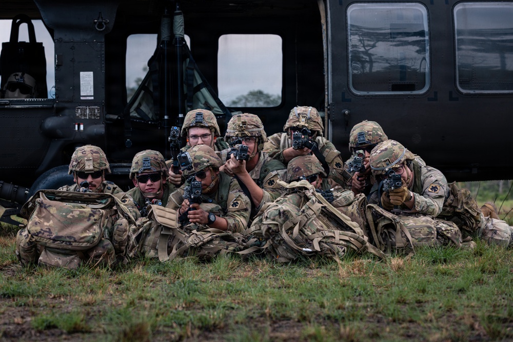 A group of approximately eight soldiers in full combat gear are positioned in a defensive perimeter on a grassy field in front of a military helicopter, likely a UH-60 Black Hawk. The soldiers are wearing MultiCam (OCP) uniforms, tactical helmets with night vision mounts, and are aiming their weapons outward in a security posture. This appears to be a training exercise or operational air assault insertion.