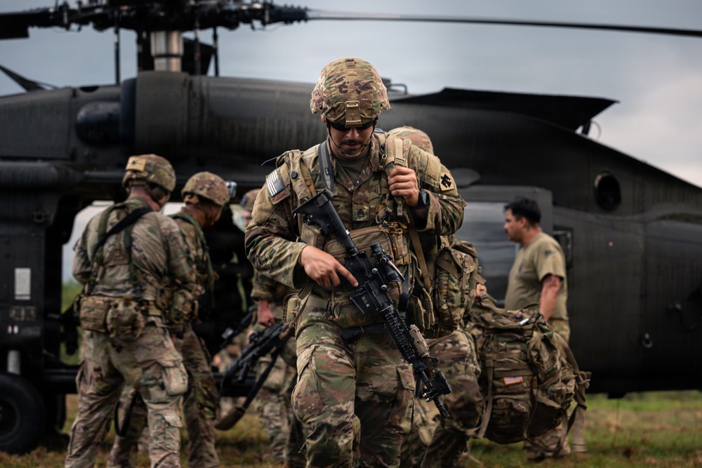 The image shows a group of military personnel in full combat gear disembarking from a dark military helicopter, likely a UH-60 Black Hawk. The lead soldier is prominently featured carrying an assault rifle and wearing multicam camouflage with a tactical helmet. The scene appears to depict a training exercise or operational insertion onto an open field.