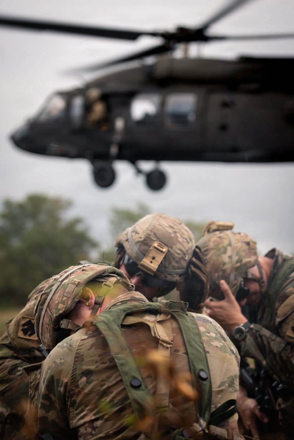 Military personnel in MultiCam/OCP camouflage uniforms are crouching and shielding themselves from rotor wash as a UH-60 Black Hawk helicopter hovers low overhead. The soldiers appear to be on a landing zone during what looks like an air assault or MEDEVAC training exercise. The foreground subjects are in sharp focus while the helicopter in the background is slightly out of focus.