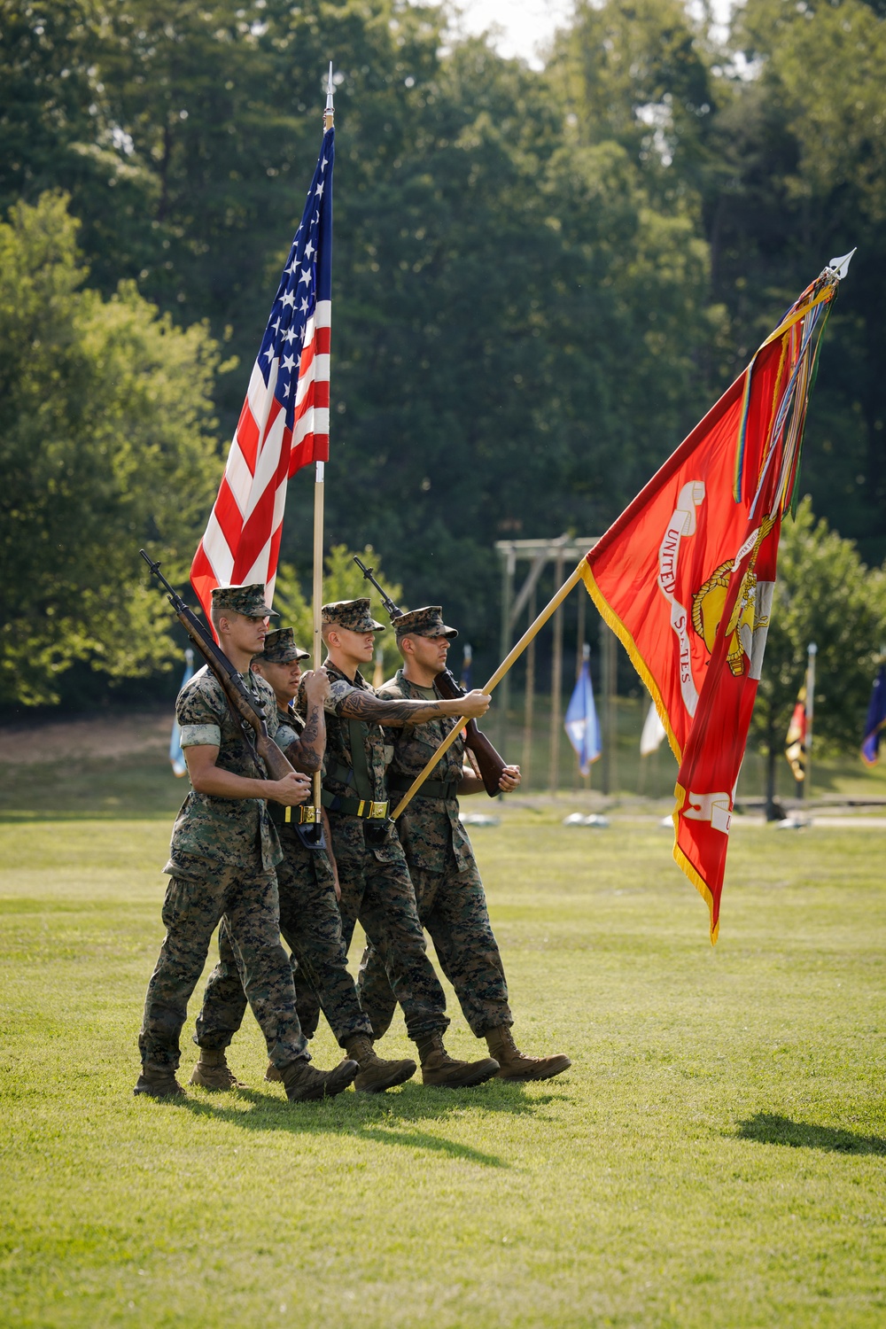 A U.S. Marine Corps color guard is marching across a grass field during a formal military ceremony. Four Marines in MARPAT camouflage uniforms carry the U.S. national flag and the Marine Corps battle color, adorned with streamers. The setting appears to be an outdoor parade ground surrounded by trees in full summer foliage.