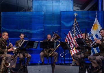 A military brass/wind ensemble of five musicians in U.S. Army uniforms performs on a stage with blue backdrop panels. Three flags are visible behind them: the United States flag, what appears to be the Uruguayan flag, and the Connecticut state flag. The formal setting suggests an official diplomatic or ceremonial event.