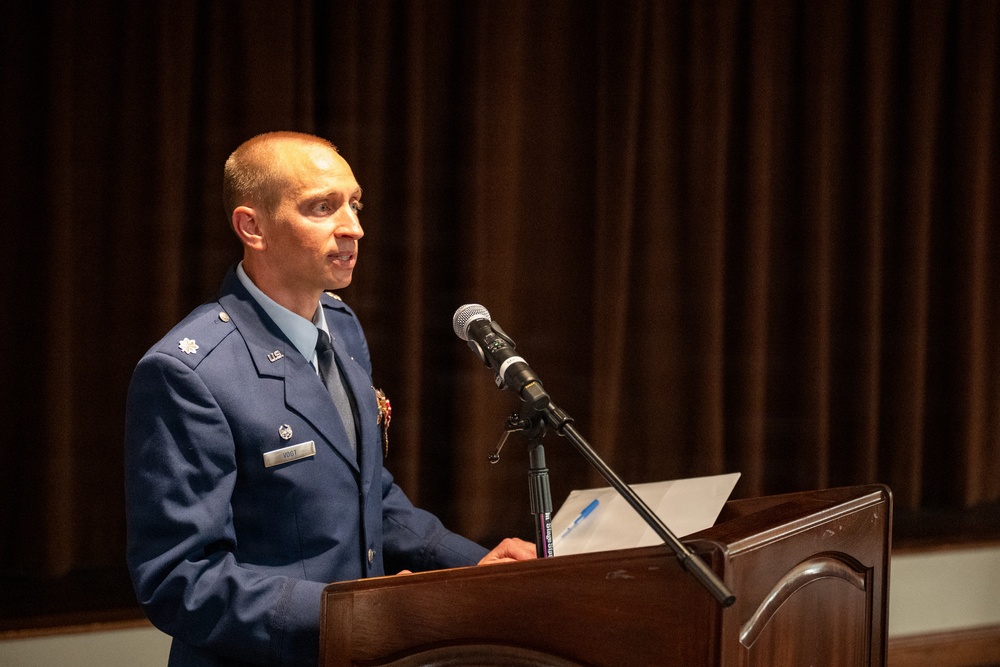 A U.S. Air Force officer in dress blue uniform is speaking at a wooden podium equipped with a microphone. The officer appears to be delivering a formal address or speech at an official military ceremony or event. Dark curtains form the backdrop of what appears to be an auditorium or formal venue.