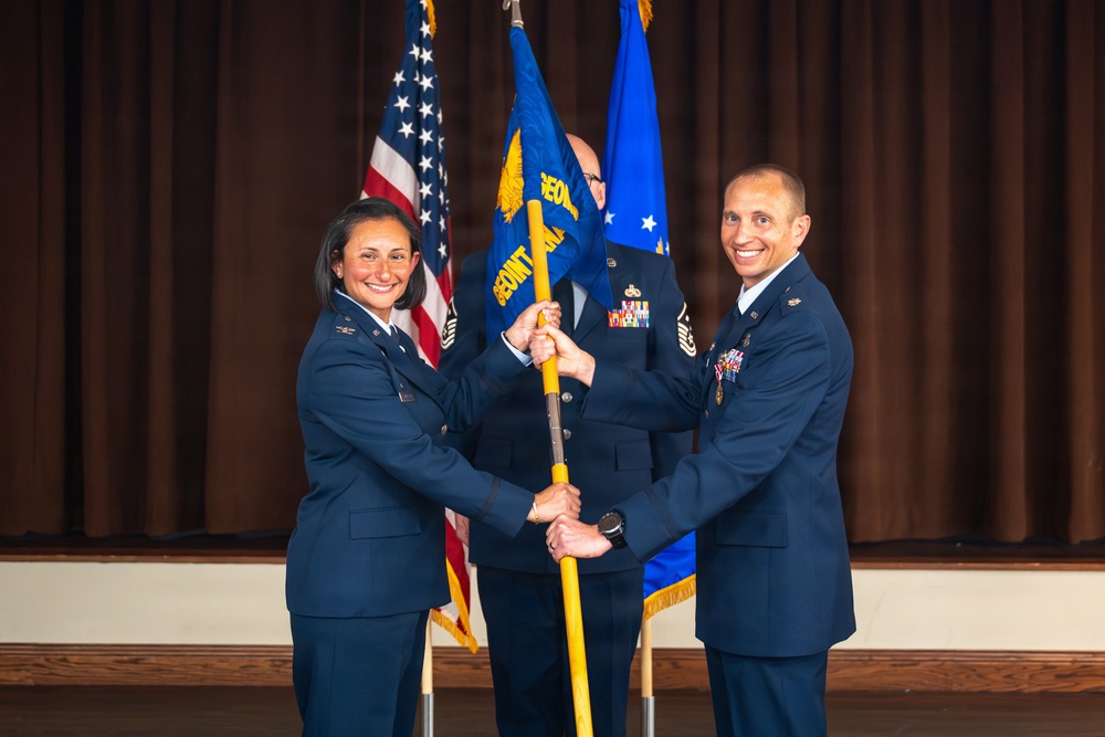 A military change of command or assumption of command ceremony is taking place, with two uniformed U.S. Air Force officers jointly holding a unit guidon (flag on a staff). A third uniformed individual stands in the background holding additional flags. Both foreground officers are smiling, indicating a formal but celebratory occasion.
