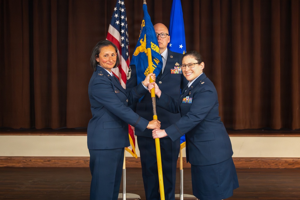 A military change of command ceremony is taking place, with two female Air Force officers jointly holding a unit guidon (flag on a pole) in a formal transfer of command. A male senior non-commissioned officer stands in the background holding the unit's blue flag. The ceremony appears to be conducted indoors on a stage with the American flag and an Air Force flag visible.